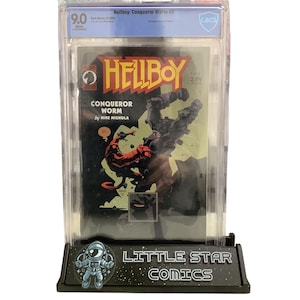 Puede incluir: Un cómic de Hellboy, "Conqueror Worm #2", calificado con 9.0, en una funda protectora. La portada muestra a Hellboy en una escena de batalla, con el título en amarillo. El cómic se exhibe en un soporte negro con el texto "LITTLE STAR COMICS".