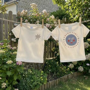 Op de afbeelding: Twee crèmekleurige t-shirts hangen aan een waslijn. Eén shirt heeft een sterontwerp, de andere een cirkelvormig ontwerp met een gezicht en de woorden "Predictably Unpredictable". De shirts zijn vastgezet met houten wasknijpers.
