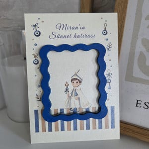 May include: A rectangular card featuring a blue wavy frame, a central illustration of a child in traditional attire holding a staff, and the text Miran'in Sünnet hatırası. The card is decorated with small blue evil eye motifs and vertical stripes.