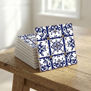 May include: A stack of square ceramic coasters with a white background and a blue and yellow floral design. The coasters are displayed on a wooden surface, with a window in the background.
