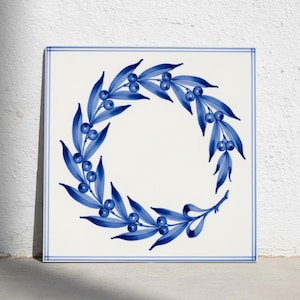 May include: A square ceramic tile featuring a white background with a hand-painted blue wreath design composed of elongated leaves and small round berries. The tile is framed by a thin blue double-line border along all four edges of the square.