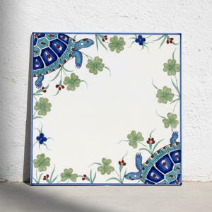 Op de afbeelding: Een vierkante keramische tegel met een witte achtergrond en twee blauwe en groenblauwe schildpadden in de tegenoverliggende hoeken. Het ontwerp bevat groene klavertjesvier, kleine rode bessen en blauwe bloemen langs de randen.