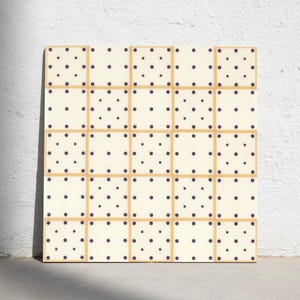 May include: A square tile featuring a grid of twenty-five smaller squares. Each small square is cream-coloured with nine dark purple polka dots arranged in a three-by-three pattern. The grid is outlined by thin, light tan lines separating each square.