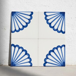 Puede incluir: Un conjunto de cuatro azulejos cuadrados de cerámica dispuestos en cuadrícula. Cada azulejo presenta un fondo blanco con un patrón en forma de abanico pintado a mano en azul en una esquina. Juntos, forman un diseño simétrico mayor.