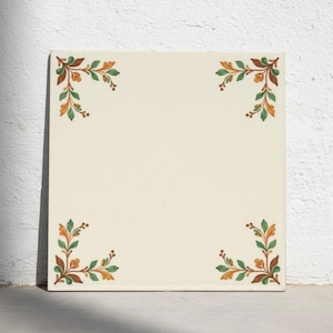 Puede incluir: Un azulejo de cerámica cuadrado con una base de color blanco roto. Cada una de las cuatro esquinas presenta un motivo botánico pintado que consiste en pequeñas hojas verdes y tallos de color marrón anaranjado en un diseño simétrico.