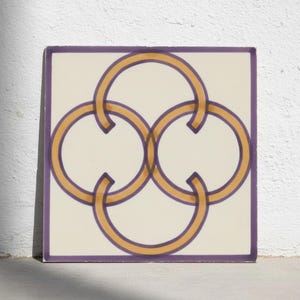 Puede incluir: Un azulejo cuadrado de cerámica con fondo blanco roto y un patrón geométrico de cuatro círculos entrelazados. Los círculos están delineados con un borde morado oscuro y un relleno amarillo dorado, creando un diseño simétrico y repetitivo.