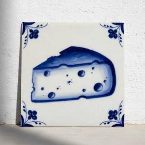 Op de afbeelding: Vierkante witte keramische tegel met een blauwe illustratie van een stuk kaas en blauwe bloemenhoekontwerpen. De kaaswig is geschilderd in een aquarel stijl.