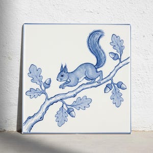 May include: A square ceramic tile featuring a blue and white illustration of a squirrel perched on an oak branch. The design includes detailed oak leaves and several acorns scattered along the branch, all rendered in a classic blue ink style.