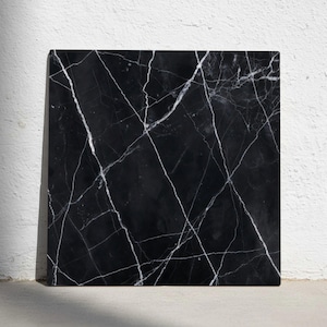 Może przedstawiać: Kwadratowa czarna płyta marmurowa z białym żyłkowaniem. Polerowana powierzchnia odbija światło, podkreślając naturalne wzory. Kamień jest ustawiony na białej ścianie, prezentując swój ciemny kolor i unikalną fakturę.