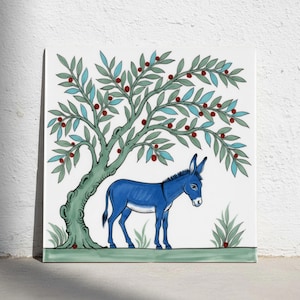 Könnte beinhalten: Eine quadratische Keramikfliese mit einer handgemalten Illustration eines blauen Esels, der unter einem stilisierten Baum mit grünen Blättern und kleinen roten Beeren steht. Die Szene ist vor einem weißen Hintergrund mit grünen Bodendetails.
