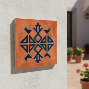 Può includere: Piastrella quadrata in terracotta con un motivo geometrico blu navy. Il design presenta triangoli, diamanti e altre forme disposte in modo simmetrico. La piastrella è montata su un muro bianco.