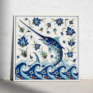 Könnte beinhalten: Eine quadratische Keramikfliese mit einem handbemalten blauen Schwertfisch, der aus stilisierten blauen Wellen springt. Der weiße Hintergrund ist mit komplexen blauen und braunen Blumenmotiven verziert, die das Fischmotiv einrahmen.