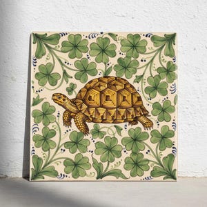 May include: A square ceramic tile featuring a central illustration of a yellow and brown tortoise. The background is decorated with a repeating pattern of green four-leaf clovers and delicate dark blue vine accents on a cream-coloured surface.