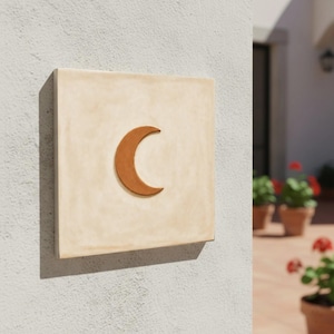 May include: A square, cream-coloured tile featuring a crescent moon. The moon is a warm brown hue and is centrally positioned. The tile is mounted on a textured, light grey wall, suitable for outdoor use.