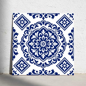 Könnte beinhalten: Quadratische Keramikfliese mit weißem Hintergrund und detailliertem blauem Blumenmuster. Das Design zeigt ein zentrales Blumenmotiv, umgeben von verzierten Schnörkeln und rautenförmigen Akzenten, was einen klassischen, dekorativen Look erzeugt.