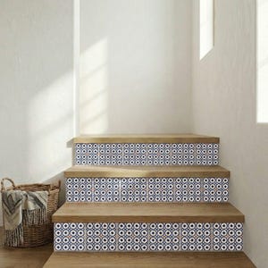 Op de afbeelding: Houten trap met decoratieve tegelstootborden. De tegels hebben een herhalend patroon van blauwe, witte en oranje cirkels. De treden zijn van lichtgekleurd hout. Een rieten mand met een deken staat ernaast.