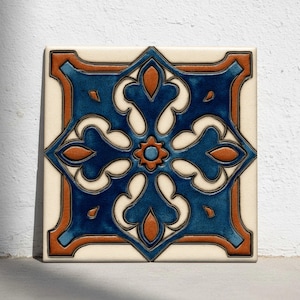 Op de afbeelding: Vierkante keramische tegel met een symmetrisch bloemmotief in blauw en oranje. De tegel heeft een crèmekleurige achtergrond en een donkeroranje rand. Het centrale ontwerp is een stervormig patroon met een kleine oranje bloem in het midden.