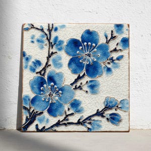 Puede incluir: Azulejo cerámico cuadrado con un diseño floral azul. Las flores y ramas están pintadas en tonos azules con detalles dorados, sobre un fondo blanco texturizado. El azulejo tiene un aspecto rústico y envejecido.