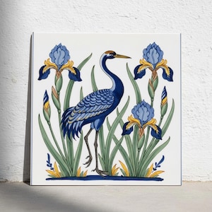 May include: A square ceramic tile featuring a hand-painted illustration of a blue crane standing among blue and yellow iris flowers with green leaves. The design is set against a white background with a thin blue border framing the entire square tile.