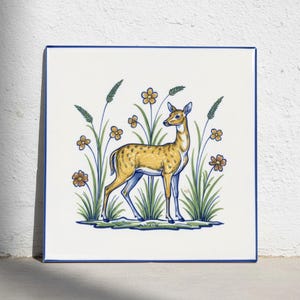 Puede incluir: Un azulejo de cerámica cuadrado con fondo blanco y borde azul. El centro presenta una ilustración pintada a mano de un ciervo de color tostado de pie entre hierba verde, tallos altos y flores naranjas con centros azules, delineado en azul.
