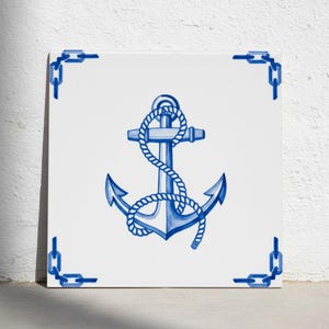 Op de afbeelding: Een vierkante witte keramische tegel met een centraal blauw geschilderd anker omwikkeld met een touw. Het ontwerp bevat een blauw kettingpatroon in elk van de vier hoeken van de tegel, wat een samenhangend nautisch visueel element vormt.