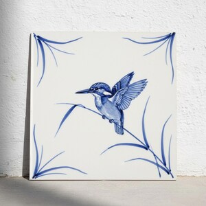 Op de afbeelding: Een vierkante keramische tegel met een blauw-witte, met de hand geschilderde illustratie van een ijsvogel die op een dun rietje zit. Het ontwerp bevat delicate blauwe hoekaccenten en een witte achtergrond, wat een klassiek vogelmotief toont.