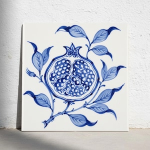 Puede incluir: Un azulejo de cerámica cuadrado con un diseño pintado a mano en azul y blanco de una granada cortada por la mitad. La ilustración muestra las semillas interiores y la corona de la fruta, rodeada de ramas con hojas en azul cobalto clásico.