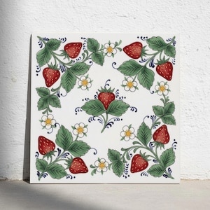 May include: A square ceramic tile featuring a white background with a repeating pattern of red strawberries, green leaves, and small white flowers with yellow centres. The design is arranged symmetrically with a central strawberry motif.