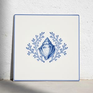 May include: A square white ceramic tile featuring a blue border and a central illustration of a seashell flanked by two symmetrical coral branches. The design is rendered in a detailed blue line art style against a plain white background.