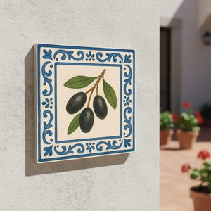 Może przedstawiać: Kwadratowa płytka ceramiczna przedstawiająca gałązkę oliwek. Oliwki są czarne, z zielonymi liśćmi, na kremowym tle. Płytka ma niebieską dekoracyjną obwódkę z kwiatowymi i zwijanymi wzorami. Płytka jest zamontowana na białej ścianie.