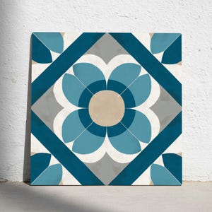 Puede incluir: Azulejo cuadrado con un diseño floral en tonos azules, grises y beige. La flor central está rodeada por un borde en forma de diamante y detalles en las esquinas. El azulejo está apoyado contra una pared blanca.