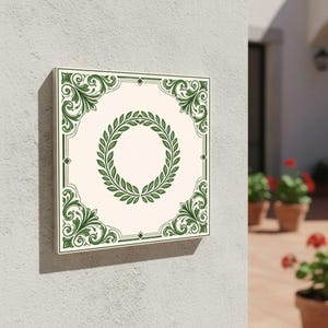 May include: Square wall art featuring a cream-colored background with a green laurel wreath in the center. The border and corners have ornate green scrollwork. The art is mounted on a white wall.