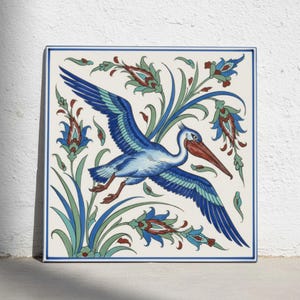 Könnte beinhalten: Eine quadratische Keramikfliese mit einer zentralen Abbildung eines fliegenden blau-weißen Pelikans. Der Vogel hat einen langen orangefarbenen Schnabel und ist von stilisierten Blumenmotiven in Blau-, Grün- und Rottönen auf cremefarbenem Grund umgeben.