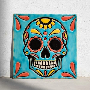 Könnte beinhalten: Quadratische Keramikfliese mit einem farbenfrohen Sugar-Skull-Design. Der Schädel ist vor einem türkisfarbenen Hintergrund platziert und mit orangefarbenen, gelben und schwarzen Details verziert. Die Fliese hat eine glänzende Oberfläche und einen schwarzen Rand.
