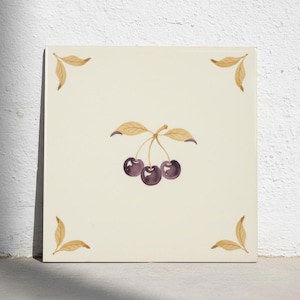 May include: A square off-white ceramic tile featuring a central illustration of three dark purple cherries with light brown stems and leaves. The four corners of the tile are decorated with matching light brown leaf motifs in a symmetrical pattern.