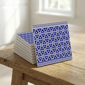 Może przedstawiać: Stos kwadratowych ceramicznych podkładek z geometrycznym wzorem niebieskich i białych gwiazd. Podkładki ułożone są na drewnianej powierzchni, z jedną podkładką ustawioną pionowo, aby zaprezentować wzór. Wzór zawiera trójkąty i gwiazdy.