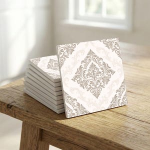 May include: A stack of square ceramic tiles with a damask pattern in shades of gray and beige. The tiles are stacked on a rustic wooden surface, with a window in the background.