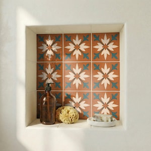 Può includere: Una nicchia del bagno con piastrelle decorative con un motivo floreale bianco, verde acqua e marrone. Una bottiglia di vetro marrone con una pompa, una spugna naturale e un piccolo portasapone bianco con sapone sono sullo scaffale.