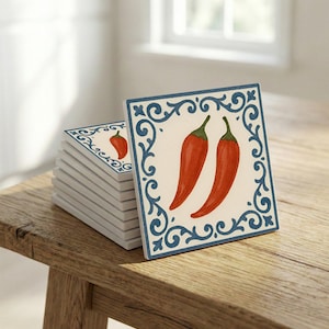 Puede incluir: Una pila de posavasos cuadrados de cerámica con un diseño de pimiento rojo. Cada posavasos presenta un fondo blanco con un borde decorativo azul. Los posavasos están apilados sobre una superficie de madera.