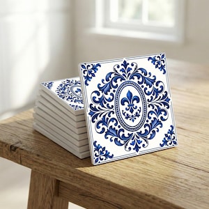 Könnte beinhalten: Ein Stapel quadratischer Keramikfliesen mit weißem Hintergrund und blauem Blumendesign. Das Design zeigt ein Oval mit einer Lilie und verzierten Schnörkeln. Die Fliesen liegen auf einer Holzoberfläche.