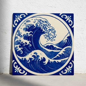 Puede incluir: Azulejo cerámico cuadrado con una ilustración azul y blanca de una gran ola oceánica, inspirada en el arte japonés. La ola está representada dentro de un marco circular, rodeada por un borde decorativo. La obra captura el movimiento dinámico del agua.