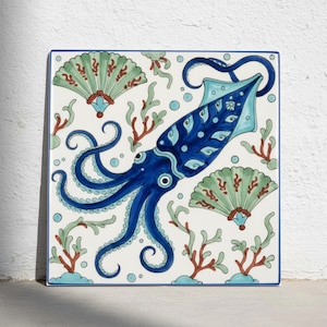 Puede incluir: Un azulejo de cerámica cuadrado que presenta un calamar azul oscuro central con detalles en azul claro, rodeado por dos conchas en forma de abanico verde claro, ramas rojas tipo coral y pequeños círculos azul claro sobre fondo blanco.