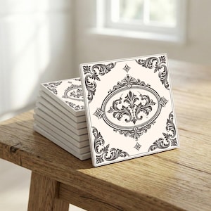 Pode incluir: Uma pilha de bases de cerâmica quadradas com um design ornamental em preto e creme. Cada base apresenta um oval central com um motivo floral, emoldurado por volutas decorativas e detalhes nos cantos. As bases estão empilhadas sobre uma superfície de madeira.