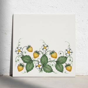 May include: A square white ceramic tile featuring a decorative illustration of strawberry plants. The design includes green leaves, white flowers with yellow centres, and yellow strawberries arranged along the bottom half of the tile.