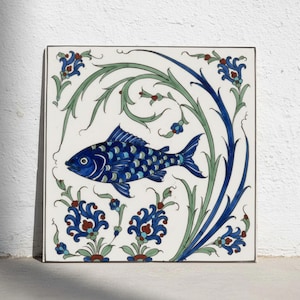 Puede incluir: Un azulejo de cerámica cuadrado con fondo blanco que presenta un pez azul central con escamas detalladas. El diseño está rodeado por enredaderas verdes fluidas y motivos florales simétricos en tonos azul, verde y rojo, creando un patrón decorativo.