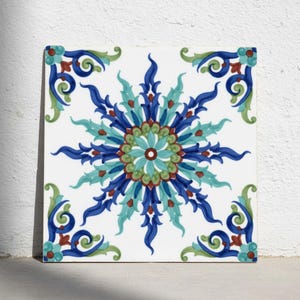 May include: A square ceramic tile featuring a central star-shaped floral motif in shades of dark blue, turquoise, and sage green. The design includes intricate swirling patterns and small red accents, with matching decorative corners on a white background.