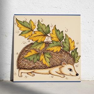 Könnte beinhalten: Eine quadratische Dekofliese mit einem braunen Igel mit cremefarbenem Gesicht und blauem Auge. Der Igel ist im Profil dargestellt und trägt ein Bündel stilisierter grüner und gelber Herbstblätter auf dem Rücken vor einem hellbeigen Hintergrund.