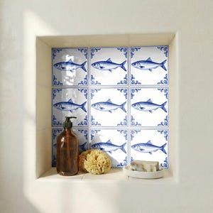 May include: Nine white ceramic tiles with a blue fish design and decorative blue borders. The tiles are arranged in a grid pattern. A brown bottle, a natural sponge, and a bar of soap are in the foreground.