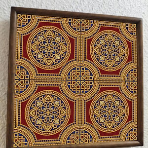 Könnte beinhalten: Quadratisches Kunstwerk in einem Holzrahmen, das ein sich wiederholendes Muster aus goldenen, blauen und roten geometrischen Formen zeigt. Das Design hat eine mosaikartige Qualität mit kreisförmigen und quadratischen Elementen. Der Rahmen ist dunkelbraun.