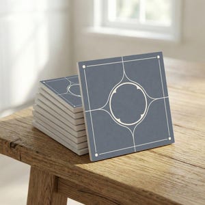 Puede incluir: Una pila de baldosas cerámicas cuadradas con un diseño geométrico. Cada baldosa es de color azul grisáceo apagado con un patrón circular y lineal blanco. Las baldosas están apiladas sobre una superficie de madera, iluminadas por luz natural.